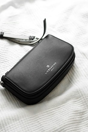 A sleek, black leather wallet or clutch with a simple, elegant design and a brand logo embossed on its surface is placed on a textured white fabric background. A detachable strap is partially visible, suggesting versatility and functionality.