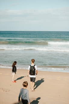 A man with a backpack walks on a sandy beach alongside two children. The children are engaged with their surroundings. The ocean waves gently hit the shore under a clear sky.