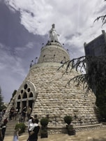 A large stone structure resembling a cone with a statue of a figure wearing a crown at its peak. The sky is partly cloudy, and people are gathered at the base of the structure, which also features arched entrances. Surrounding the area are small trees and a high-rise building in the background.