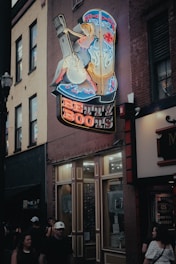 A vibrant neon sign in the shape of a large cowboy boot is mounted on the side of a building. The sign features a stylized cowgirl playing a guitar with colorful neon accents, including red, blue, and yellow lights. Below the illustration, the words 'Betty Boots' are brightly lit in red and blue. The building is part of a lively street scene with people walking by, dressed casually, and other signs indicating businesses like a whiskey store. The atmosphere suggests an urban setting, possibly a downtown or entertainment district.