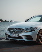 A Mercedes-Benz car is parked near a waterfront with a serene body of water in the background. The car's front grille and headlights are prominently visible, and a ship can be seen on the water far behind.