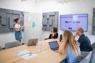 A small group of people are gathered in a modern conference room. One person stands by a whiteboard presenting a diagram labeled 'UGC Types', while the others are seated around a wooden table with laptops and papers. A large screen on the wall displays content related to the presentation.
