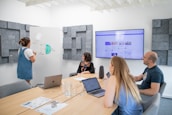 A group of four people engaged in a meeting inside a modern conference room. One person is standing and presenting at a whiteboard filled with a pie chart discussing 'UGC Types', while the others are seated at a table. Laptops and documents are in use, with a large screen displaying an online interface in the background. The room has acoustic wall panels and a neat, modern design.