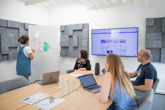 A group of four people engaged in a meeting inside a modern conference room. One person is standing and presenting at a whiteboard filled with a pie chart discussing 'UGC Types', while the others are seated at a table. Laptops and documents are in use, with a large screen displaying an online interface in the background. The room has acoustic wall panels and a neat, modern design.