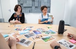 A group of people sitting around a table with various photographs spread out in front of them. One person is holding up a postcard-sized image while others are looking on. The setting is a conference room with a minimalist design and acoustic paneling on the walls. Notepads, pens, and art supplies are scattered across the table.
