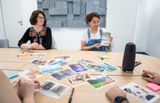 A group of people sitting around a table with various photographs spread out in front of them. One person is holding up a postcard-sized image while others are looking on. The setting is a conference room with a minimalist design and acoustic paneling on the walls. Notepads, pens, and art supplies are scattered across the table.