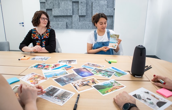 A group of people sitting around a table with various photographs spread out in front of them. One person is holding up a postcard-sized image while others are looking on. The setting is a conference room with a minimalist design and acoustic paneling on the walls. Notepads, pens, and art supplies are scattered across the table.