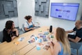 A group of four people sit around a table in a modern office setting. They are engaged in a creative discussion, examining various colorful photographs spread out on the table. A large screen on the wall displays a website or social media platform interface. The atmosphere appears collaborative and lively.