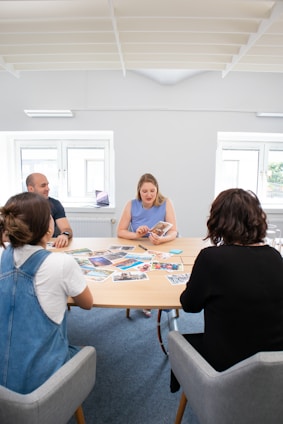 A group of four people are seated around a table with various photographs spread out on its surface. The room is well-lit with natural light coming through two large windows. The individuals appear to be engaged in a discussion or meeting.