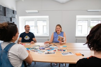 A group of four individuals is seated around a table, engaged in a discussion. The table is scattered with various photographs and there are sticky notes and markers visible. The setting appears to be a bright, well-lit room with two large windows in the background. The people seem to be engaged in a creative or planning session.