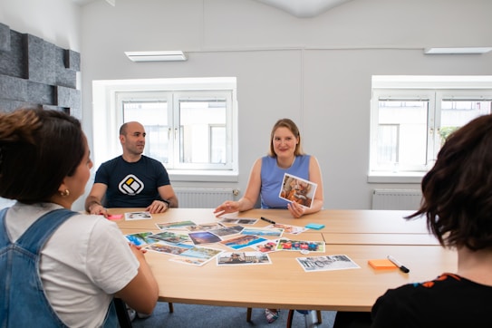 A group of four individuals is seated around a table, engaged in a discussion. The table is scattered with various photographs and there are sticky notes and markers visible. The setting appears to be a bright, well-lit room with two large windows in the background. The people seem to be engaged in a creative or planning session.