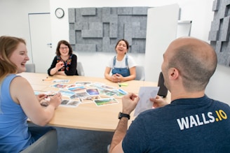 Four people are sitting around a conference table, laughing and engaging in conversation. There are various photographs spread out on the table. The setting appears casual and creative, with soundproofing panels on the wall in a light-filled room.