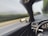 A view from the interior of a car, focusing on the dashboard and part of the steering wheel. The side mirror reflects the blurred landscape, indicating motion and speed along a road flanked by trees and greenery.