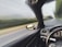 A view from the interior of a car, focusing on the dashboard and part of the steering wheel. The side mirror reflects the blurred landscape, indicating motion and speed along a road flanked by trees and greenery.