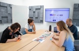 Four people sit around a wooden conference table in a modern meeting room. A large screen on the wall shows a website. The group appears to be discussing or reviewing documents and images spread out on the table. The room has acoustic panels on the walls, a whiteboard, and a speaker on the table.