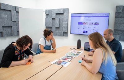 Four people sit around a wooden conference table in a modern meeting room. A large screen on the wall shows a website. The group appears to be discussing or reviewing documents and images spread out on the table. The room has acoustic panels on the walls, a whiteboard, and a speaker on the table.