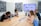 Four people sit around a wooden conference table in a modern meeting room. A large screen on the wall shows a website. The group appears to be discussing or reviewing documents and images spread out on the table. The room has acoustic panels on the walls, a whiteboard, and a speaker on the table.