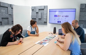 Four people sit around a wooden conference table in a modern meeting room. A large screen on the wall shows a website. The group appears to be discussing or reviewing documents and images spread out on the table. The room has acoustic panels on the walls, a whiteboard, and a speaker on the table.