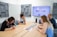 Four people sit around a wooden conference table in a modern meeting room. A large screen on the wall shows a website. The group appears to be discussing or reviewing documents and images spread out on the table. The room has acoustic panels on the walls, a whiteboard, and a speaker on the table.