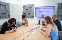 Four people sit around a wooden conference table in a modern meeting room. A large screen on the wall shows a website. The group appears to be discussing or reviewing documents and images spread out on the table. The room has acoustic panels on the walls, a whiteboard, and a speaker on the table.