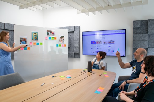 A group of four people are engaged in a presentation or meeting in a modern office setting. A woman in a blue dress stands at a whiteboard covered with colorful sticky notes and photographs, pointing towards them while other attendees, seated around a wooden table, listen and take notes. The table is scattered with pens and more sticky notes. A large screen displaying a website or digital content is mounted on the wall.