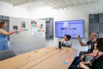 A group of four people are engaged in a presentation or meeting in a modern office setting. A woman in a blue dress stands at a whiteboard covered with colorful sticky notes and photographs, pointing towards them while other attendees, seated around a wooden table, listen and take notes. The table is scattered with pens and more sticky notes. A large screen displaying a website or digital content is mounted on the wall.