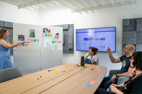 A group of four people are engaged in a presentation or meeting in a modern office setting. A woman in a blue dress stands at a whiteboard covered with colorful sticky notes and photographs, pointing towards them while other attendees, seated around a wooden table, listen and take notes. The table is scattered with pens and more sticky notes. A large screen displaying a website or digital content is mounted on the wall.