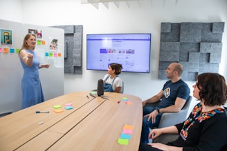 A meeting or presentation in a modern office setting, where a woman in a blue dress is standing in front of a whiteboard with colorful sticky notes. Three other people are seated around a wooden conference table, focused on a large wall-mounted screen displaying a website or social media page. The room has gray acoustic paneling on the walls.