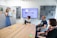 A meeting or presentation in a modern office setting, where a woman in a blue dress is standing in front of a whiteboard with colorful sticky notes. Three other people are seated around a wooden conference table, focused on a large wall-mounted screen displaying a website or social media page. The room has gray acoustic paneling on the walls.