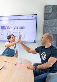 Two people are sitting at a wooden table in a well-lit room, giving each other a high five. The person on the left is wearing blue overalls and a white shirt, while the person on the right is wearing a black t-shirt with a logo. In the background, a large screen displays a webpage with the hashtag #LoveWallsio.