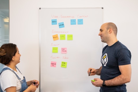 Two people stand in front of a whiteboard covered with colorful sticky notes. The notes are arranged into categories: 'User-Generated Content,' 'Engagement,' 'Event,' and 'Social Wall.' The individuals appear to be in discussion, with one holding a marker.