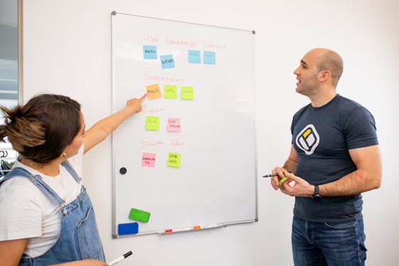 Two individuals are standing next to a whiteboard covered with colorful sticky notes. The woman on the left is pointing at a note while holding a marker, and the man on the right is also holding a marker. The sticky notes have various headings like 'User-Generated Content', 'Engagement', 'Event', and 'Social Wall', each with related subpoints underneath.