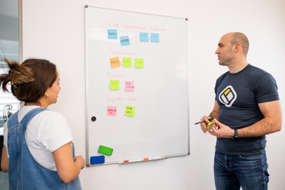 Two people engaged in a discussion in front of a whiteboard. The whiteboard has sticky notes with various categories like 'User-Generated Content' and 'Engagement', each with subcategories noted beneath them. One person holds a marker and an eraser, while the other listens attentively.