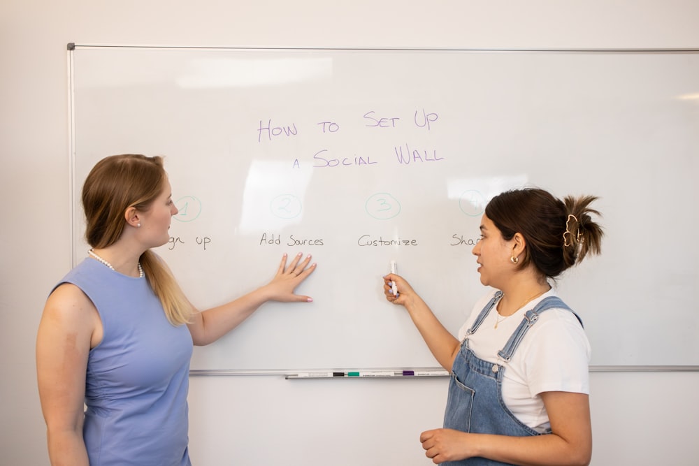 two women standing in front of a whiteboard with writing on it
