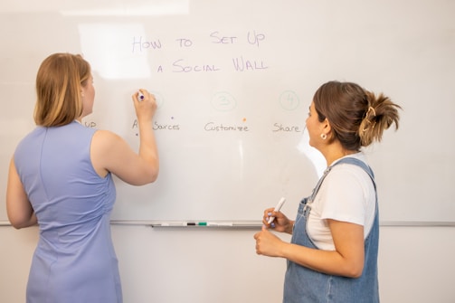 Two people are writing on a whiteboard, discussing the setup process for a social wall. The board is divided into sections labeled: setup, add sources, customize, and share. One person is wearing a blue dress and the other is wearing denim overalls.