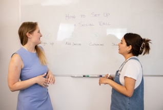 Two people are standing in front of a whiteboard discussing a topic titled 'How to Set Up a Social Wall'. The whiteboard outlines steps: Sign up, Add Sources, Customize, and Share. One person is holding a marker, and both are engaged in conversation, likely explaining the steps.