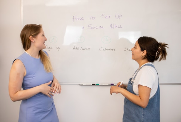 Two people are standing in front of a whiteboard discussing a topic titled 'How to Set Up a Social Wall'. The whiteboard outlines steps: Sign up, Add Sources, Customize, and Share. One person is holding a marker, and both are engaged in conversation, likely explaining the steps.