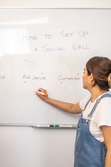 A woman wearing denim overalls stands in front of a whiteboard and writes with an orange marker. The whiteboard has writing that explains how to set up a 'Social Wall,' with steps numbered one to three.