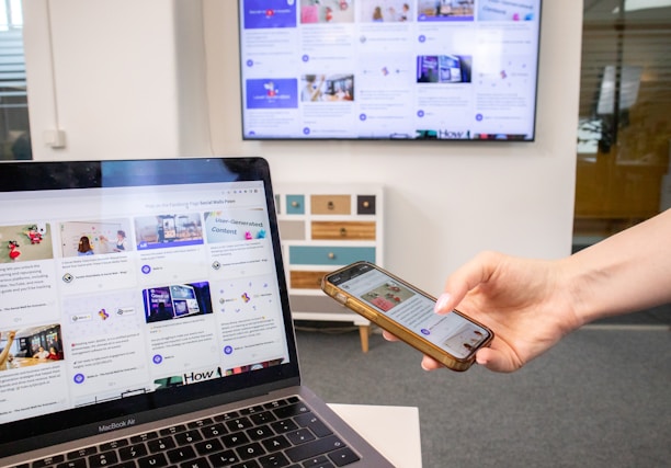 A hand holds a smartphone displaying a webpage, while a laptop, also showing similar content, is in the foreground. In the background, a large monitor on the wall is displaying multiple web pages or a content grid. The setting appears to be an office with a grey carpet and a white and multicolored cabinet.