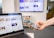 A hand holds a smartphone displaying a webpage, while a laptop, also showing similar content, is in the foreground. In the background, a large monitor on the wall is displaying multiple web pages or a content grid. The setting appears to be an office with a grey carpet and a white and multicolored cabinet.