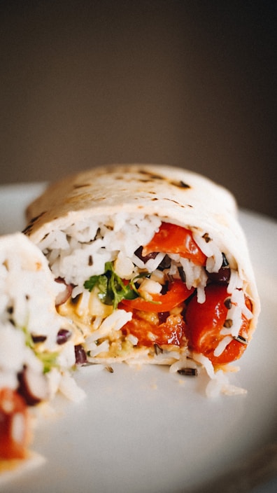 A close-up view of a burrito cut in half, displaying its colorful filling. Inside the burrito, there is visible white rice, fresh greens, red bell peppers, and purple beans, all wrapped in a tortilla. The ingredients appear fresh and vibrant, and the tortilla has a slight char.