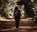 A person wearing a red bandana and a backpack walks along a forest path. The pathway is covered with dry leaves, and dense green foliage surrounds the trail, creating a serene and natural setting. The sun filters through the trees, casting dappled shadows on the ground.