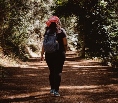 A person wearing a red bandana and a backpack walks along a forest path. The pathway is covered with dry leaves, and dense green foliage surrounds the trail, creating a serene and natural setting. The sun filters through the trees, casting dappled shadows on the ground.