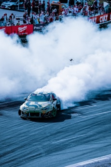 A sports car is drifting on a race track, producing a large cloud of smoke from its tires. A crowd of spectators is visible in the background behind barriers, watching the action. The scene conveys high energy and excitement typical of motorsport events.