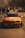 An orange modified sports car with various logos and decals on the bonnet and windshield is parked on a road. A crowd of people can be seen in the background, possibly at a car event or a racing venue. The setting appears to be outdoors with a mix of sunlight and shadows. Another car is visible in the background, to the right.