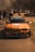 An orange modified sports car with various logos and decals on the bonnet and windshield is parked on a road. A crowd of people can be seen in the background, possibly at a car event or a racing venue. The setting appears to be outdoors with a mix of sunlight and shadows. Another car is visible in the background, to the right.