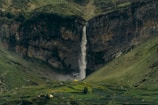 A waterfall cascades down a rugged cliff surrounded by lush, green hills. At the base of the waterfall, there are hints of mist and vegetation thriving amidst the rocky terrain. A solitary yellow tent is visible in the lower region, adding a splash of color and suggesting a sense of adventure or exploration in the serene landscape.