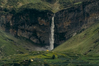 A waterfall cascades down a rugged cliff surrounded by lush, green hills. At the base of the waterfall, there are hints of mist and vegetation thriving amidst the rocky terrain. A solitary yellow tent is visible in the lower region, adding a splash of color and suggesting a sense of adventure or exploration in the serene landscape.