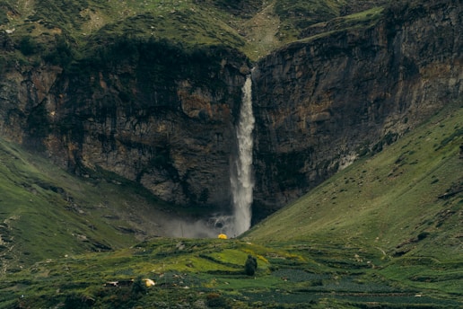 A waterfall cascades down a rugged cliff surrounded by lush, green hills. At the base of the waterfall, there are hints of mist and vegetation thriving amidst the rocky terrain. A solitary yellow tent is visible in the lower region, adding a splash of color and suggesting a sense of adventure or exploration in the serene landscape.