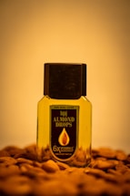 A bottle of Bajaj Almond Drops hair oil is placed on a surface scattered with almonds. The bottle is transparent with a black cap, showing the golden-colored oil inside. The label on the bottle emphasizes '6X Vitamin E' nourishment.